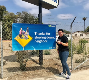 Person standing next to a sign on a fence, holding a thumbs-up, outdoors on a sunny day.